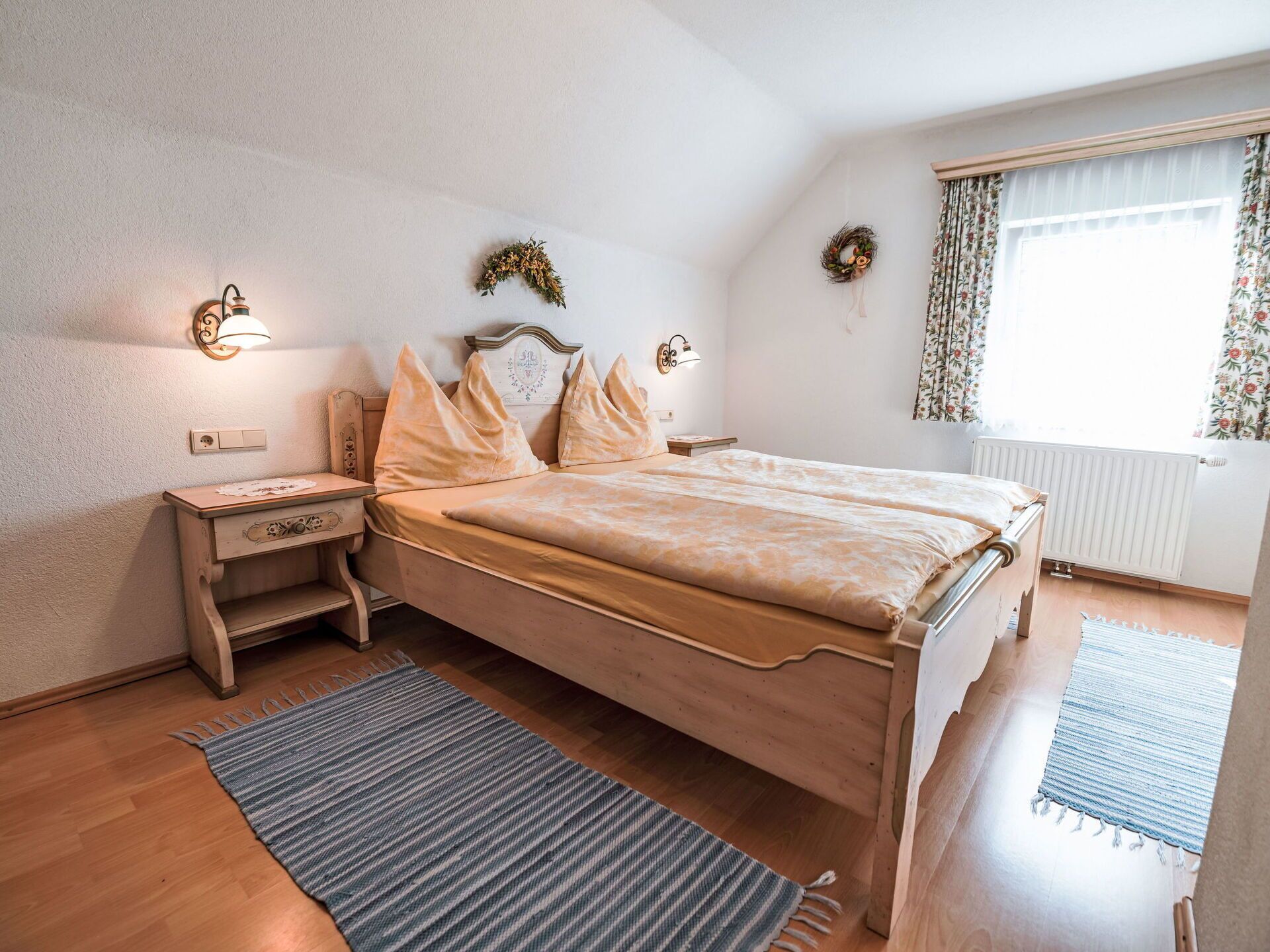 A bedroom with a wooden bed, two nightstands, a blue striped rug, and a window with floral curtains.