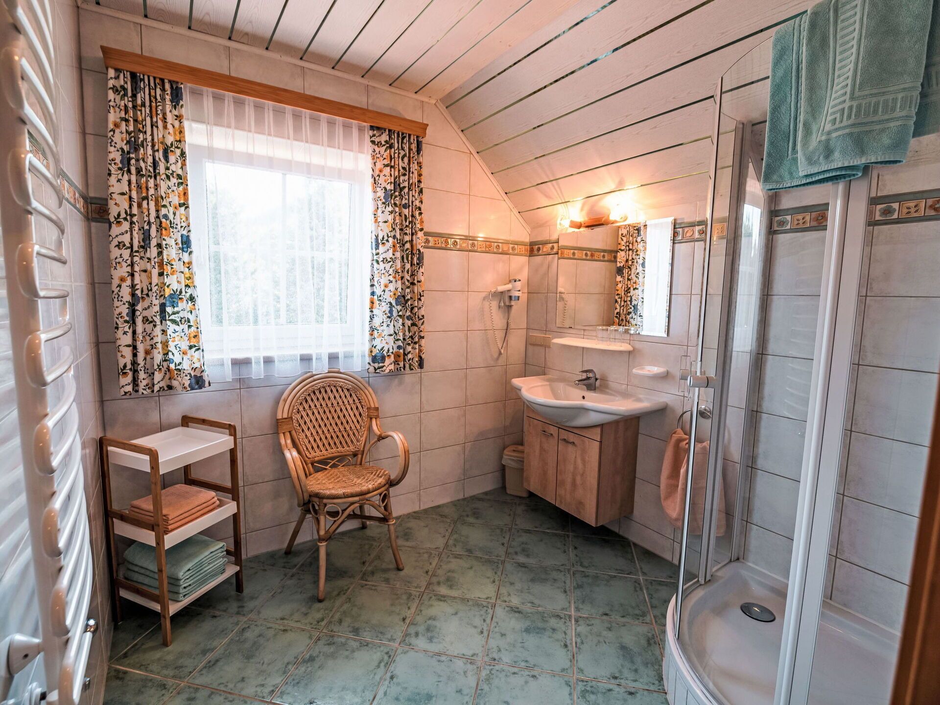 A bathroom with a shower, a sink, a chair, and a window with floral curtains.