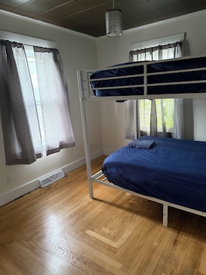 Un lit superposé avec un matelas bleu, une structure en métal blanc, une fenêtre avec des rideaux, un plancher en bois et un luminaire suspendu.