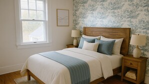 Un dormitorio con una cama de madera, dos mesitas de noche con lámparas, un ventanal y un papel tapiz con motivos florales.