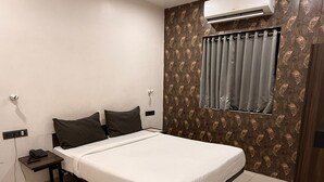 A bedroom with a bed, a nightstand, an air conditioning unit, a window with curtains, and a wall with a patterned wallpaper. - HOTEL LOTUS (Surat)