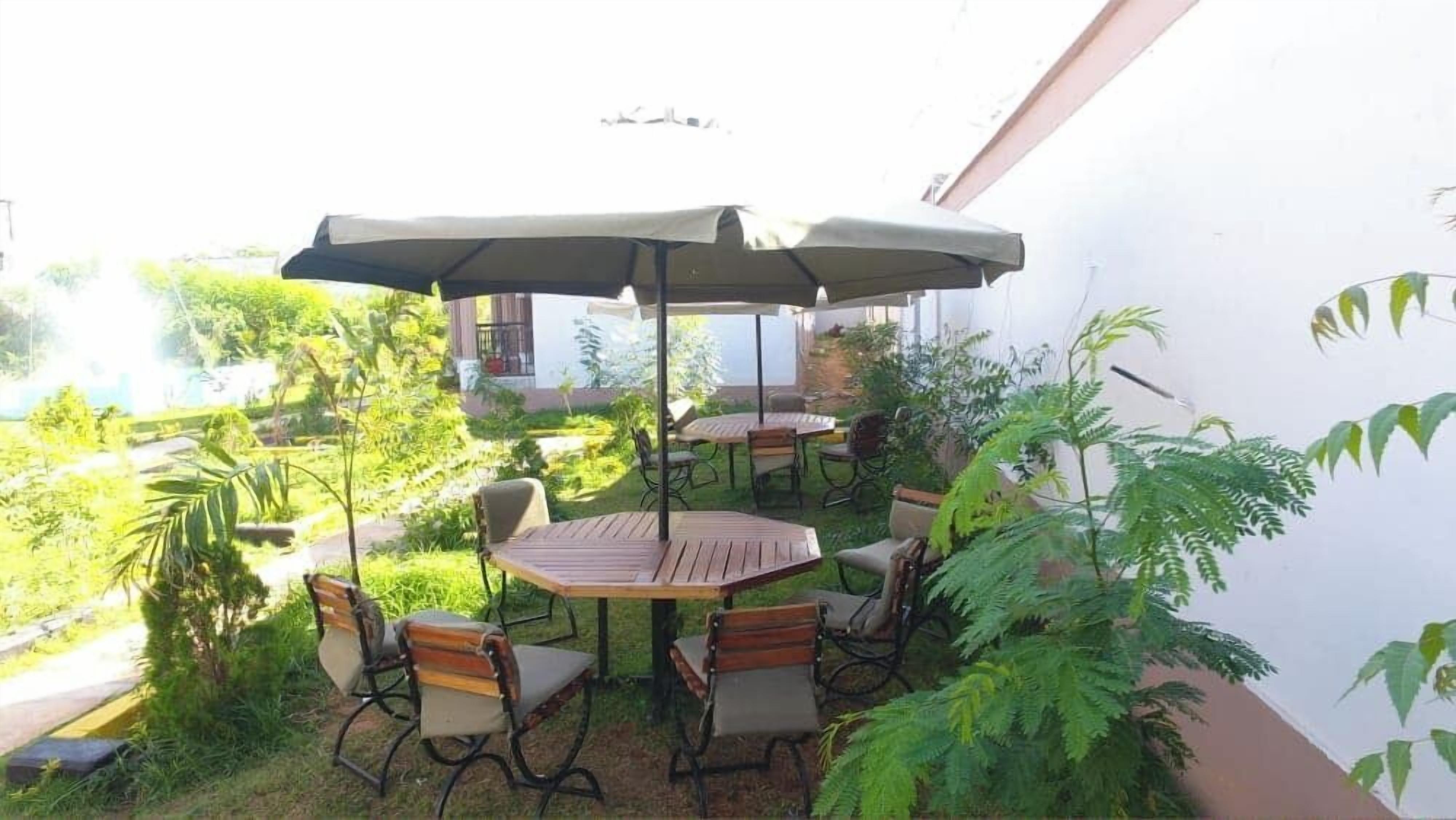 Un patio al aire libre con mesas y sillas de madera, un paraguas grande y abundante vegetación.