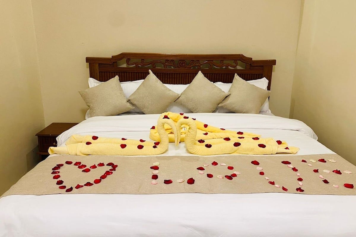 A neatly made bed with a heart-shaped arrangement of rose petals and a towel folded in the shape of a heart.