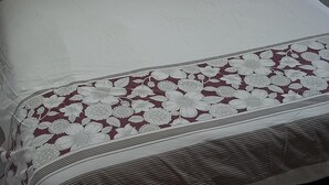 Sängyssä on kukkakuviollinen päiväpeite ja tyynyjä, lattia on laatoitettu, ja oikealla puolella on pieni vaaleanpunainen leluauto.