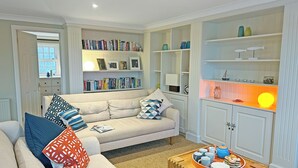 A living room with a beige sofa, a coffee table with a tray of snacks, and a bookshelf filled with books and decorative items. - Reverse-level retreat in St Mawes, perfect for family or friends. (St Mawes, near Falmouth)