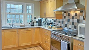 A kitchen with wooden cabinets, a stainless steel range hood, a tiled backsplash, and a window with a view of trees. - Reverse-level retreat in St Mawes, perfect for family or friends. (St Mawes, near Falmouth)