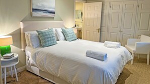 A neatly made bed with white linens and two patterned pillows, a green lamp on a bedside table, a white chair, and a large mirror above a vanity. - Reverse-level retreat in St Mawes, perfect for family or friends. (St Mawes, near Falmouth)