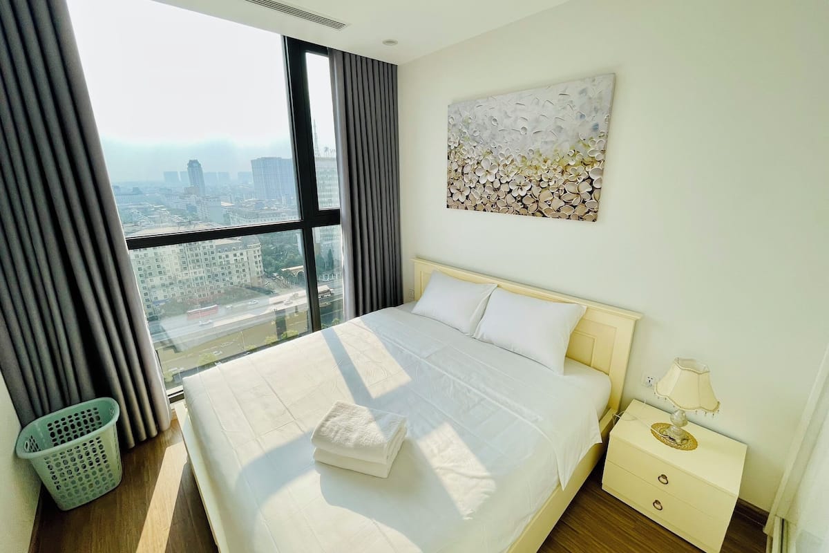 Habitación de hotel con una cama grande, una mesita de noche, una lámpara, una obra de arte en la pared y vistas de la ciudad a través de amplios ventanales.
