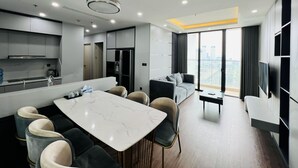 A modern kitchen with a marble countertop, a dining area with a table and chairs, a sofa, and a large window with a city view. - Hopo Apt - Vinhomes Sky Lake Luxury  (Hanoi)