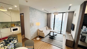 A modern hotel room with a kitchen, a living area with a sofa and coffee table, and a large window with a city view. - Hopo Apt - Vinhomes Sky Lake Luxury  (Hanoi)