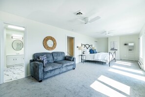 A bedroom with a bed, a gray sofa, a ceiling fan, a round mirror, and a white wardrobe. - Bayfront Escape in Edgewater with Deck, Balcony, and Walk to Beach (Edgewater)