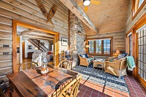 A log cabin interior with a dining area, a stone wall, wicker furniture, and a view through large windows. - Waterfront Log Home Near Sister Bay- Game Room, Kayaks & Sunrises (Sister Bay)