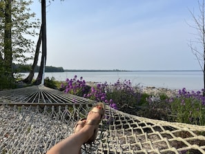 A hammock with a person's foot resting on it, overlooking a body of water with purple flowers in the foreground. - Waterfront Log Home Near Sister Bay- Game Room, Kayaks & Sunrises (Sister Bay)