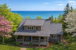 A two-story log cabin with a porch, surrounded by trees and a view of the ocean. - Waterfront Log Home Near Sister Bay- Game Room, Kayaks & Sunrises (Sister Bay)