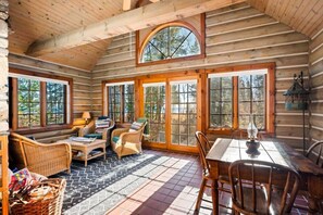 A log cabin interior with wicker furniture, a dining table, and large windows. - Waterfront Log Home Near Sister Bay- Game Room, Kayaks & Sunrises (Sister Bay)