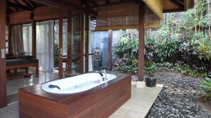A bathroom with a large wooden bathtub, a bench, and a window overlooking a garden with various plants. - Dea Villas - Villa Radha by Elite Havens (Canggu)