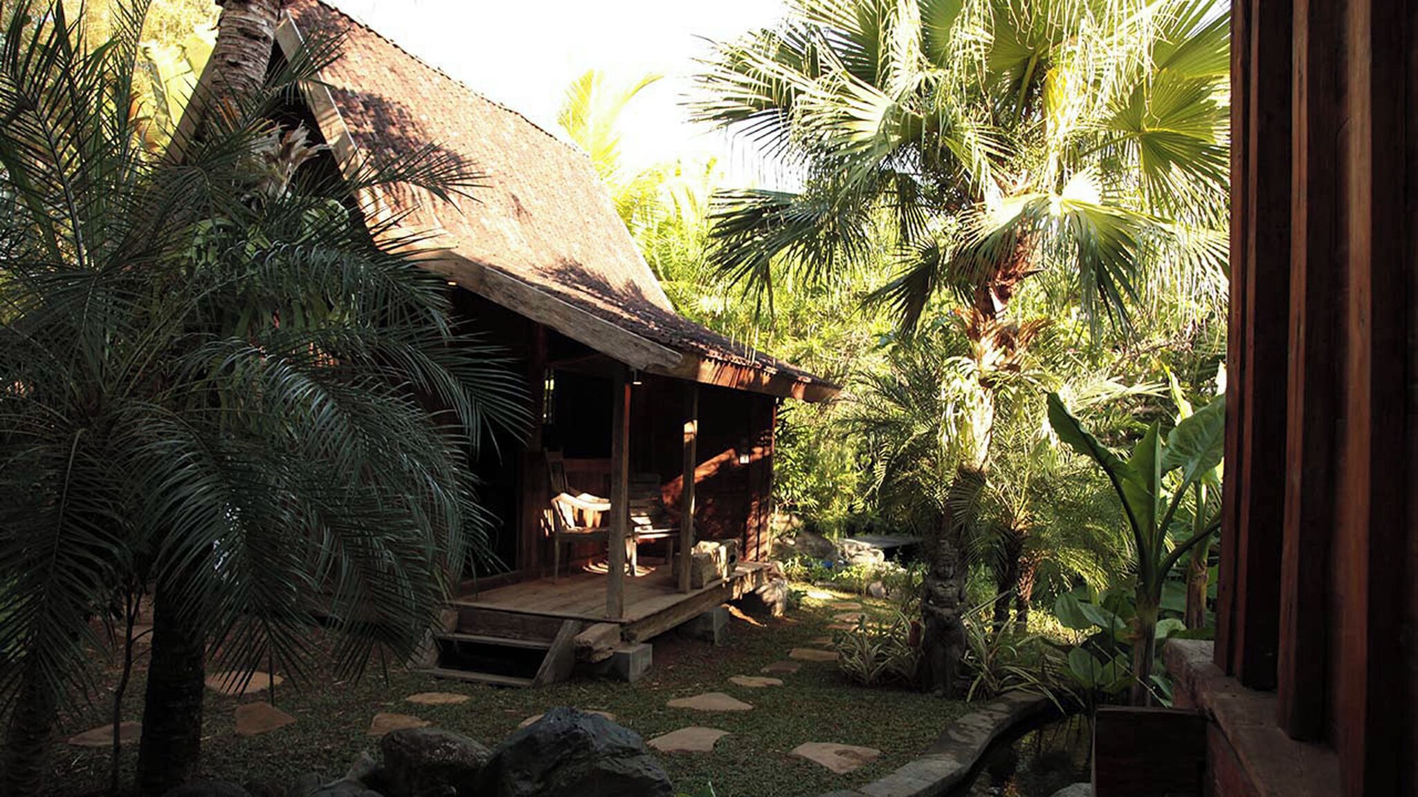 A rustic wooden structure with a thatched roof, surrounded by lush tropical vegetation including palm trees and a variety of plants.