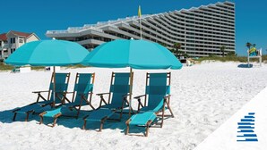 Una playa con cuatro sillas y sombrillas turquesas, un moderno edificio de hotel al fondo y un cielo azul despejado.