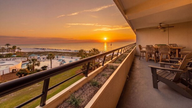 Balcón con vista al atardecer sobre el océano, zona de piscina y mobiliario exterior.