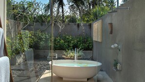 Ein modernes Badezimmer im Freien mit einer freistehenden Badewanne, umgeben von tropischen Pflanzen und einer Betonwand.
