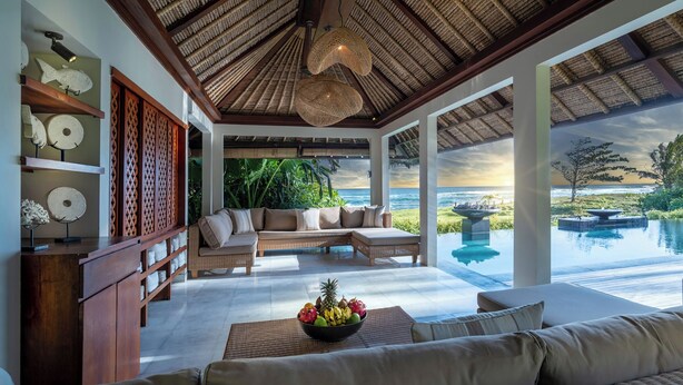Ein geräumiger Wohnbereich mit einer großen Couch, einem Esstisch und einer Obstschale. Große Fenster bieten Blick aufs Meer.