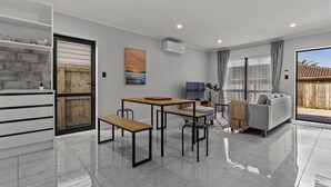 A modern living room with a sofa, a dining table with benches, a TV, and a large window with curtains. - Modern Townhouse with Parking nr Airport & Manukau (Auckland South)