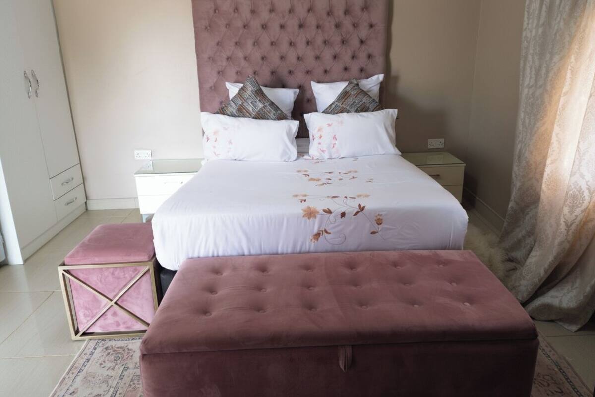 A bedroom with a large bed, a tufted headboard, two bedside tables, a pink ottoman, and a patterned rug.