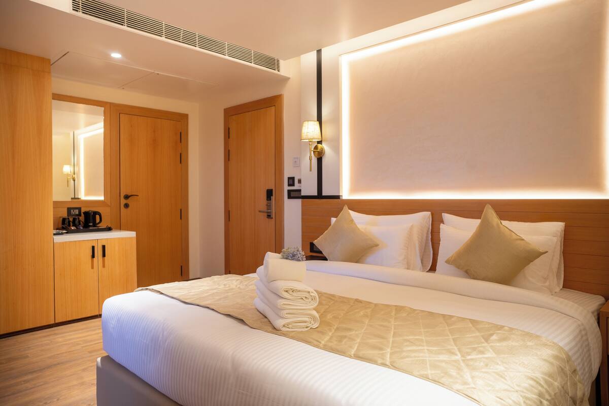 A hotel room with a neatly made bed, a wooden wardrobe, a bedside table with a lamp, and a mirror on the wall.