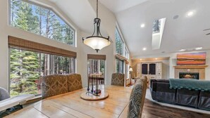 A spacious room with a large dining table, comfortable seating, a fireplace, and large windows offering a view of the forest. - Tahoe Trailside Lodge: Hot Tub, AC, Walk-to-Trail (South Lake Tahoe)