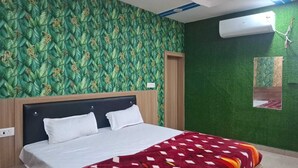 Quarto de hotel com cama, papel de parede com padrão de folhas verdes, cabeceira de madeira, colcha com padrão vermelho e branco e um aparelho de ar condicionado.