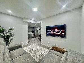 A modern living room with a grey sofa, a patterned rug, and a flat-screen TV mounted on the wall displaying a barbecue show. - Golden Retreat | Private & Modern 2BR Suite NW Calgary (Calgary)