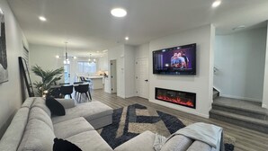 A modern living room with a sectional sofa, a flat-screen TV mounted on the wall, a fireplace, and a dining area in the background. - Family-Friendly Calgary Retreat | Gateway to Banff (Calgary)