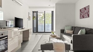 Een moderne woonkamer met een grijze bank, een kleine salontafel en een televisie. Er is een keuken met een fornuis en een koelkast.