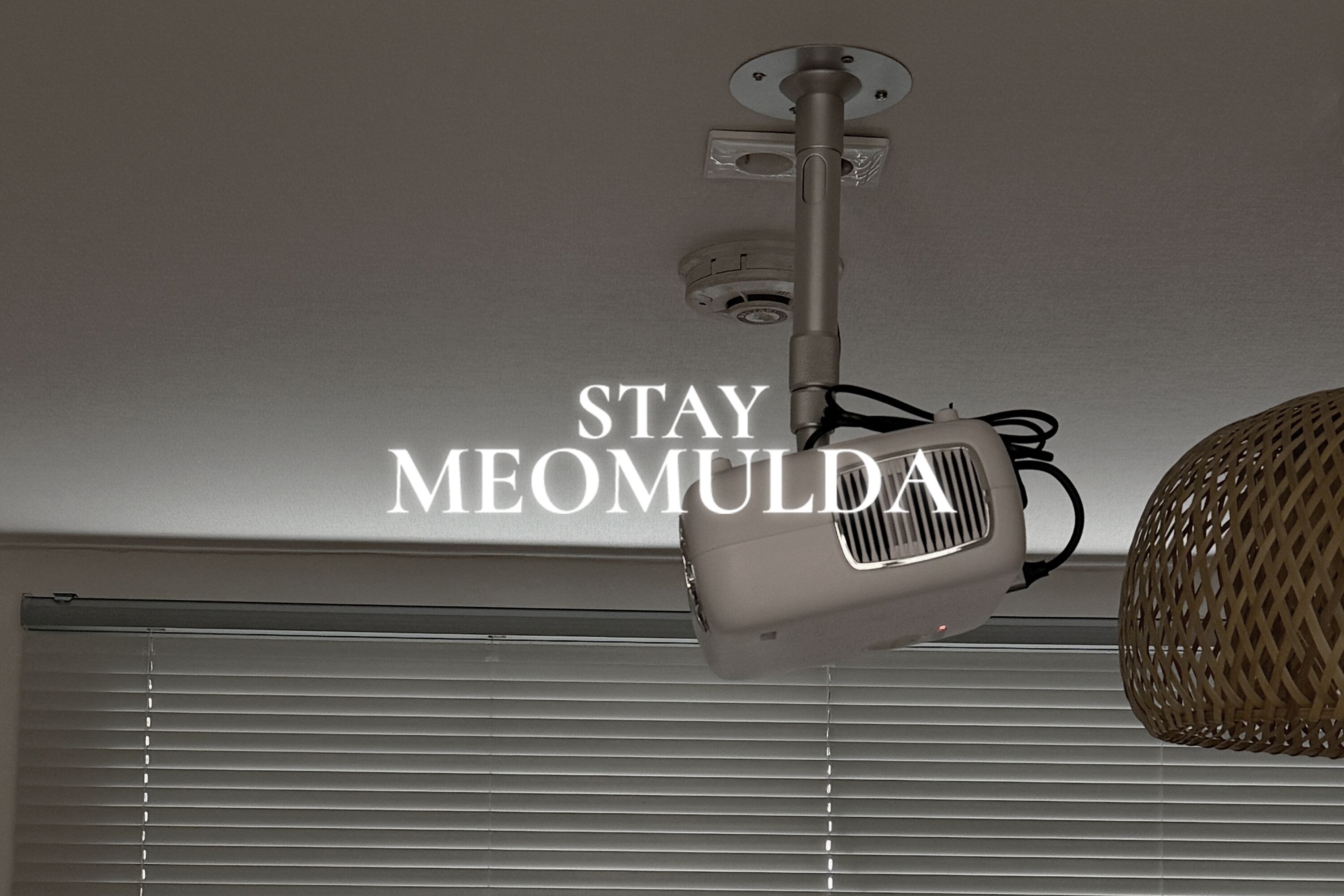 Een aan het plafond gemonteerde projector met een zichtbare stroomkabel en een label met de tekst 'STAY MEOMULDA'.