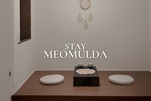 Una habitación minimalista con una mesa de madera, dos taburetes blancos y redondos, un atrapasueños en la pared y el texto "STAY MEOMULDA".
