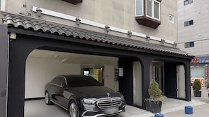 Ein schwarzes Auto parkt in einer überdachten Parkzone mit einem Gebäudeeingang, einem kleinen Topfpflanzen und zwei blauen Behältern auf dem Gehweg.