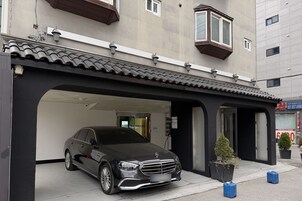 Un coche negro estacionado en una zona de estacionamiento techada, con la entrada de un edificio, una pequeña planta en maceta y dos contenedores azules en la acera.