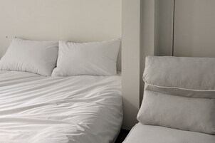 Una cama bien hecha con sábanas blancas y dos almohadas, un sillón tapizado a rayas y una pared lisa.