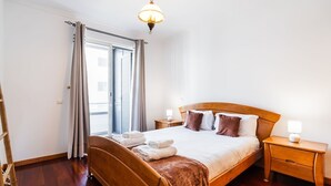 A bedroom with a wooden bed, a nightstand, a lamp, a window with curtains, and a wooden floor. - GuestReady - Charming Apartment with Ocean Views (Caniço)
