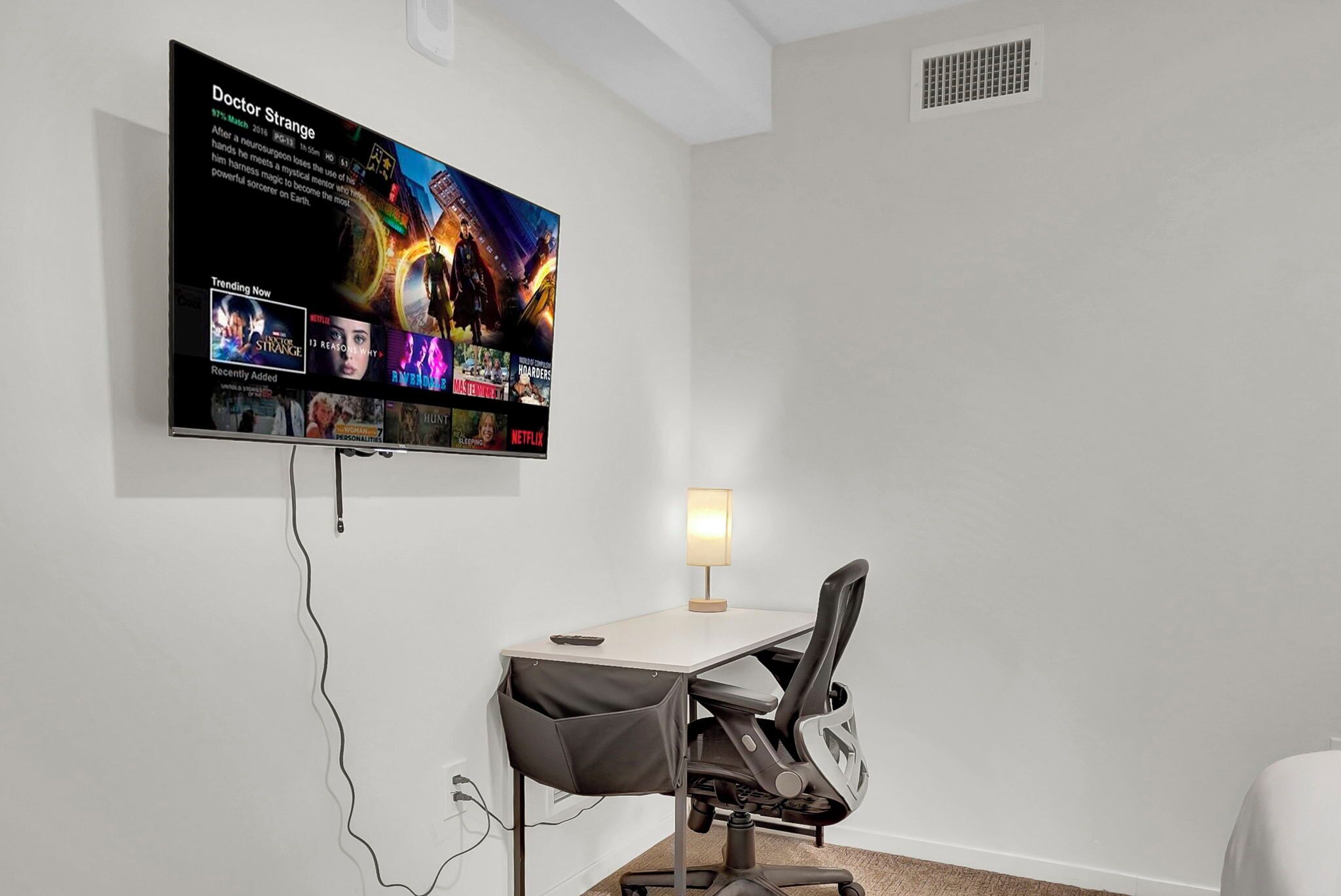 Um espaço de escritório moderno com uma televisão de tela plana grande presa na parede, exibindo um pôster de filme. Há uma mesa com uma cadeira, uma luminária e uma pequena mesa.