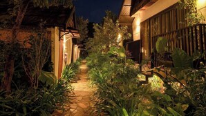 Un sentiero notturno, costeggiato da una vegetazione rigogliosa e illuminato da luci calde, conduce a un edificio dallo stile architettonico tradizionale.