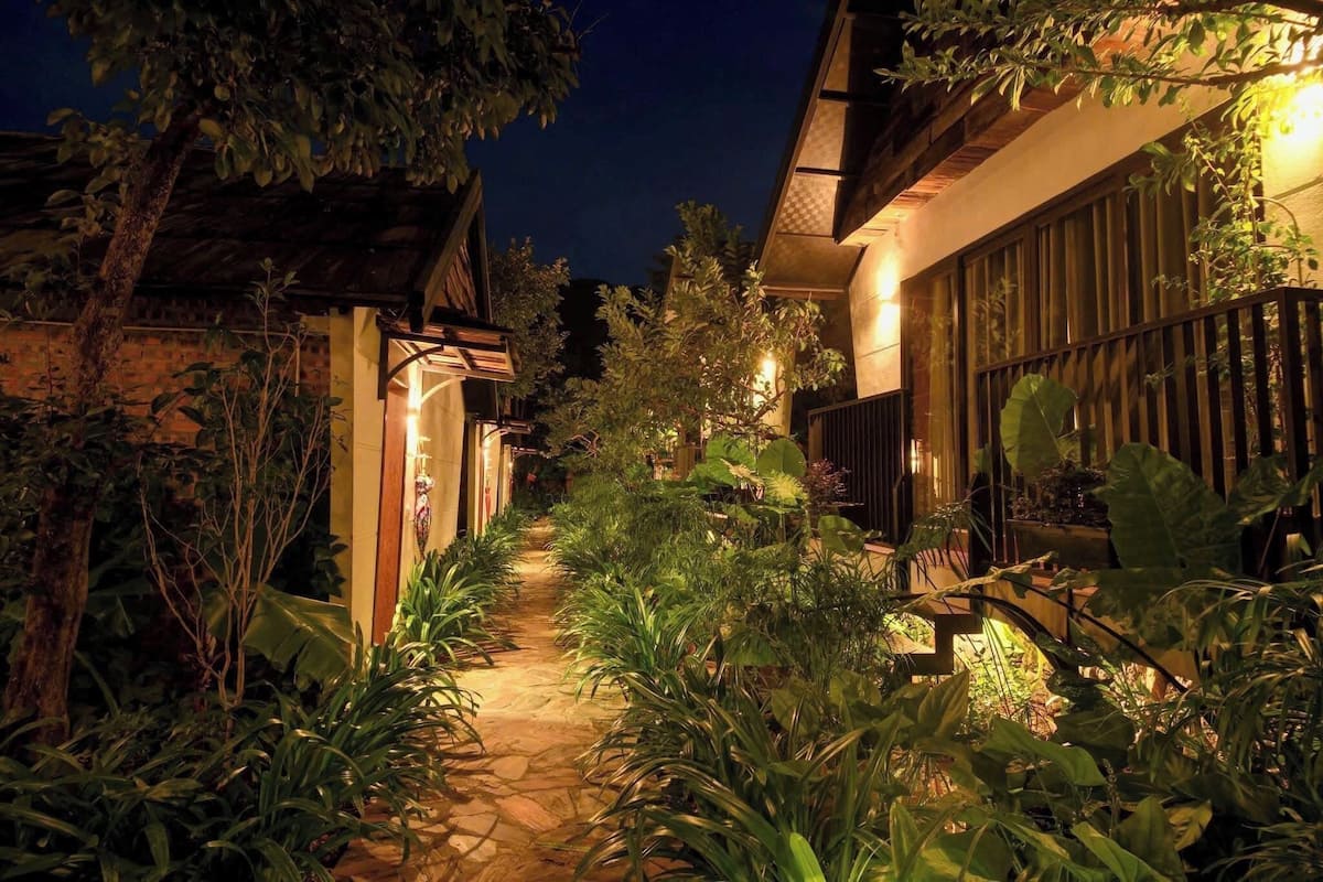 A nighttime pathway flanked by lush greenery and illuminated by warm lights, leading to a building with a traditional architectural style.