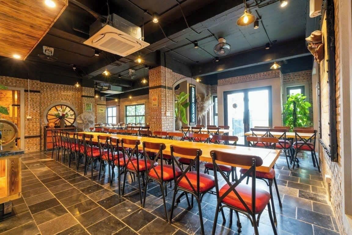 Een restaurant met lange gedeelde tafels en rode stoelen met kussens, bakstenen muren en grote ramen.