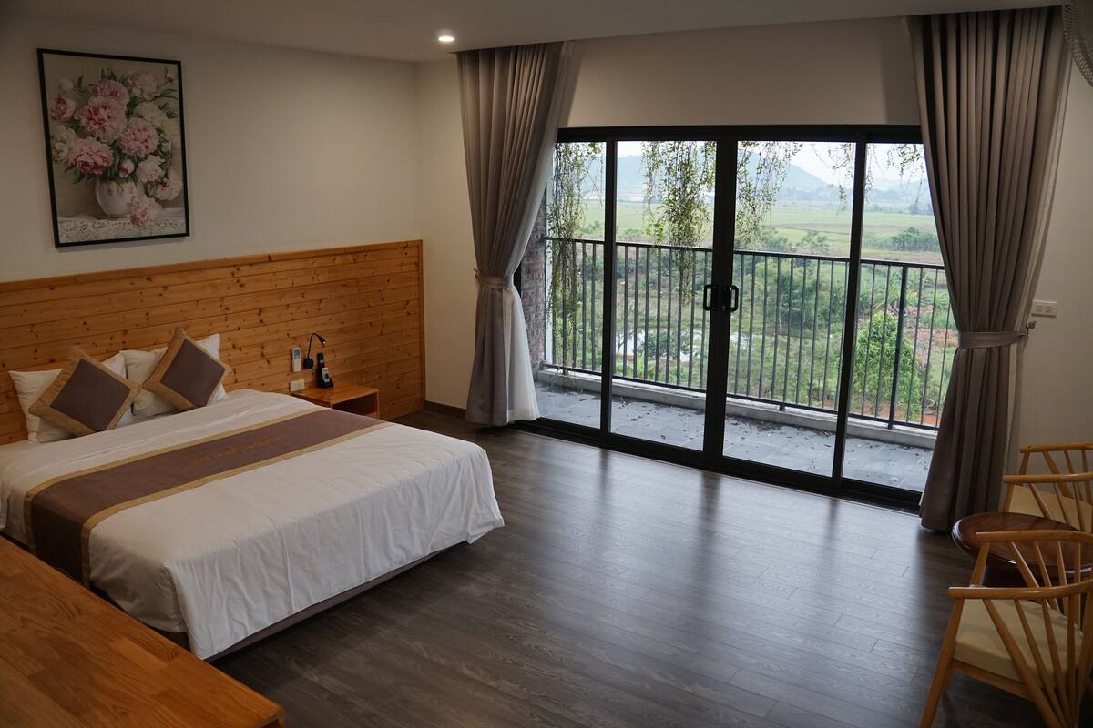 A hotel room with a large bed, wooden headboard, a wooden bench, a dining table with chairs, and a balcony with a view of greenery.