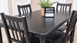 A dining table with a black tablecloth, surrounded by six dark wooden chairs. A small potted plant is placed on the table, and a sign with "WIFI PASSWORD" is visible.