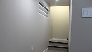A narrow hallway with light gray walls, a wooden floor, and a small window with horizontal blinds.