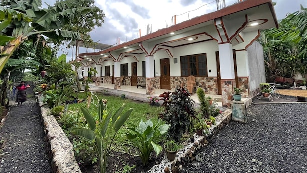 A two-story house with a covered porch, surrounded by tropical plants and a gravel driveway.