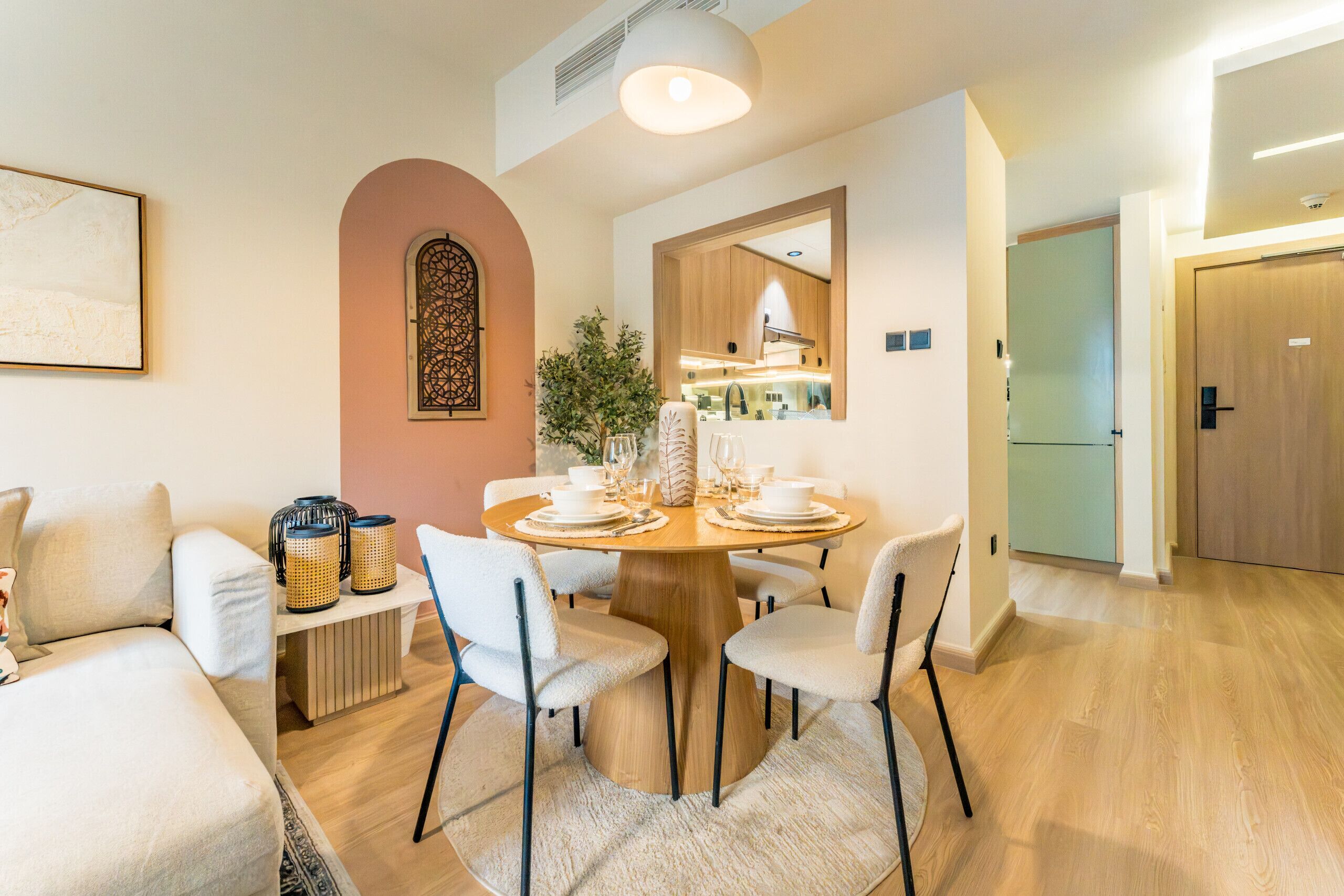 Un moderno comedor con una mesa de madera redonda dispuesta para cuatro personas, rodeada de sillas blancas. Una pequeña mesita auxiliar sostiene adornos y una planta. La habitación tiene un piso de madera clara, un gran ventanal con un marco decorativo y una puerta que conduce a otra habitación.