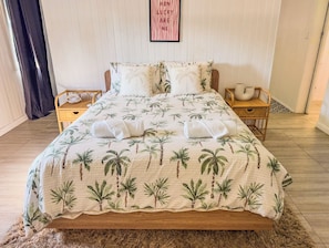 A bed with a tropical palm leaf bedspread, two pillows, a framed quote on the wall, and two nightstands with decorative items.