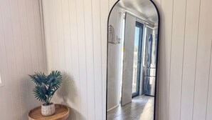 Ein Zimmer mit einem großen Spiegel, einem kleinen runden Tisch mit einer Pflanze und Blick in einen anderen Raum mit Fliesenboden.